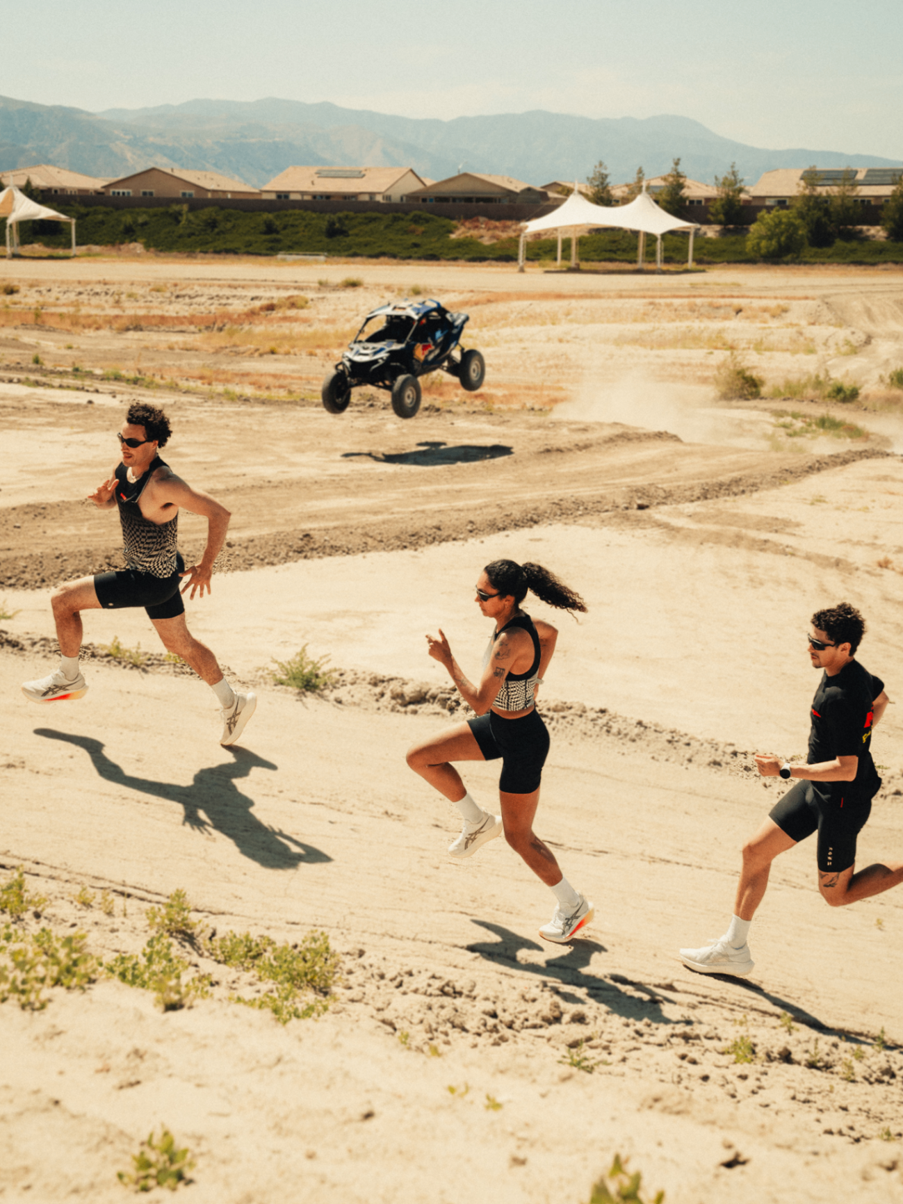 Three runners sprinting across the desert as a Red Bull Can-Am buggy launches off a jump behind them