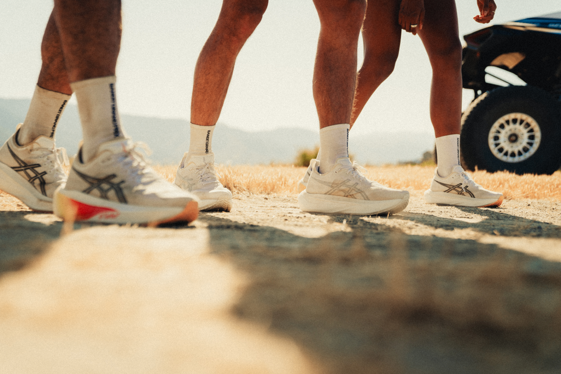 Close-up of runners' legs in Asics shoes and Bandit Grand Prix socks beside a Red Bull Can-Am buggy wheel
