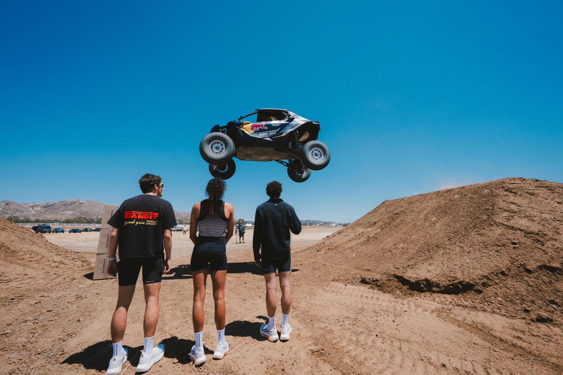 Red Bull Can-Am buggy launching off a dirt jump as three spectators in Bandit Grand Prix 2026 gear watch from below