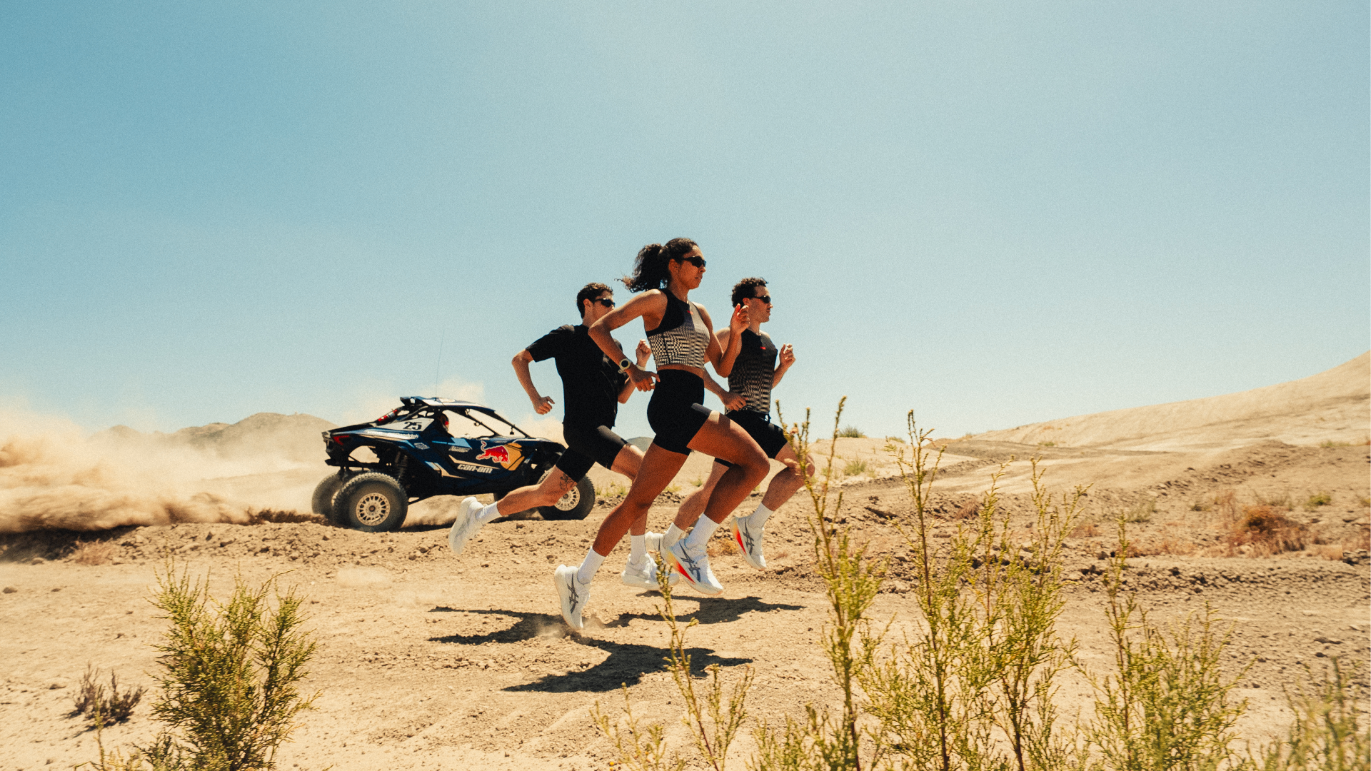 Three runners sprinting across the desert with a Red Bull Can-Am buggy kicking up dust behind